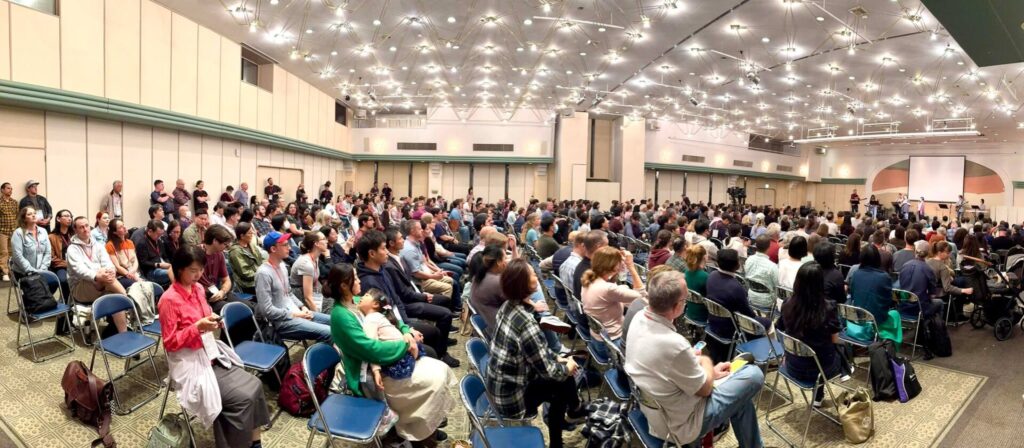 広い屋内ホールに、大勢の人々が椅子に座って前方のステージを向いている様子。天井には無数の小さなライトが輝き、会場全体が明るい。前方では数人がマイクや楽器を使って発表や演奏をしているように見える。観客は年齢や服装もさまざまで、ほぼ満席の状態。会場の後方や壁際にも立って見ている人がいて、にぎやかで集中した雰囲気が伝わる。