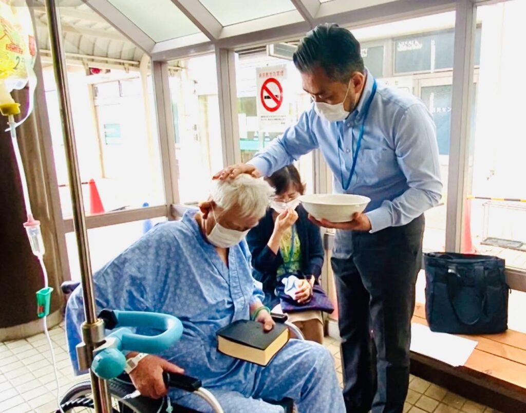 明るい施設内のスペースで、車椅子に座った高齢の男性がキリスト教の洗礼を受けている。男性は病院用の服を着てマスクを着用し、膝の上に聖書を置いている。前に立つ牧師が、器に入った水を手にし、受洗者の頭にそっと手を置いて洗礼を施している。そばには付き添いと思われる高齢の女性が座り、静かに見守っている。受洗者の体調に配慮しながら行われる、穏やかで厳かな洗礼の場面である。