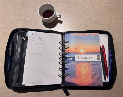 黒いファスナー付きの手帳カバーが机の上に開かれて置かれている。中にはリング式の手帳があり、左ページは日付入りのスケジュール欄、右ページは夕焼けの海を写した表紙デザインになっている。右側には赤と青のペンが差し込まれ、左側にもペンが収納されている。手帳の上方には白いカップに入った飲み物があり、全体的に落ち着いた色合いで、作業や計画の時間を感じさせる雰囲気。